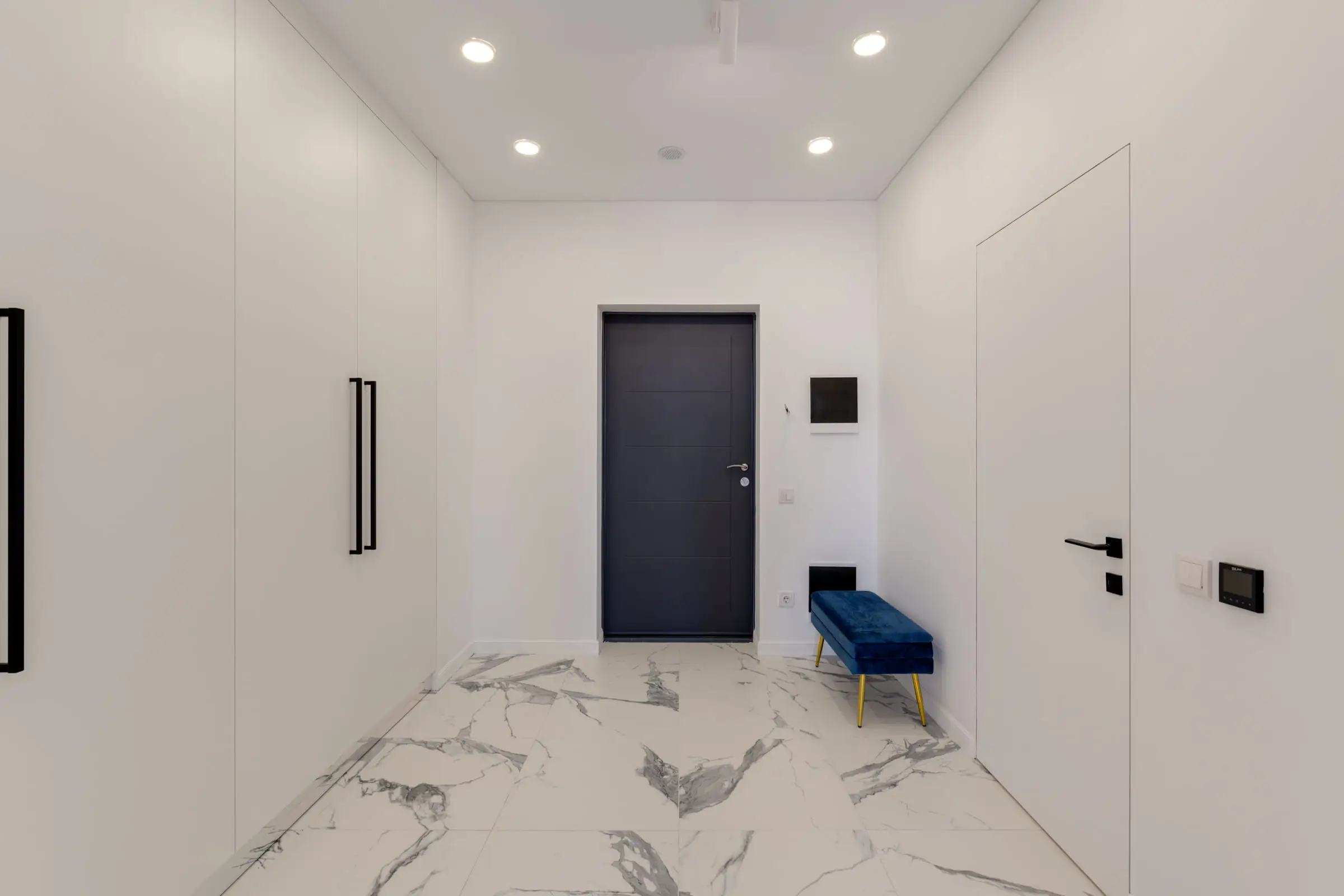Entrée d’appartement au sol de marbre blanc en grandes dalles, porte sombre, banc bas, murs blancs