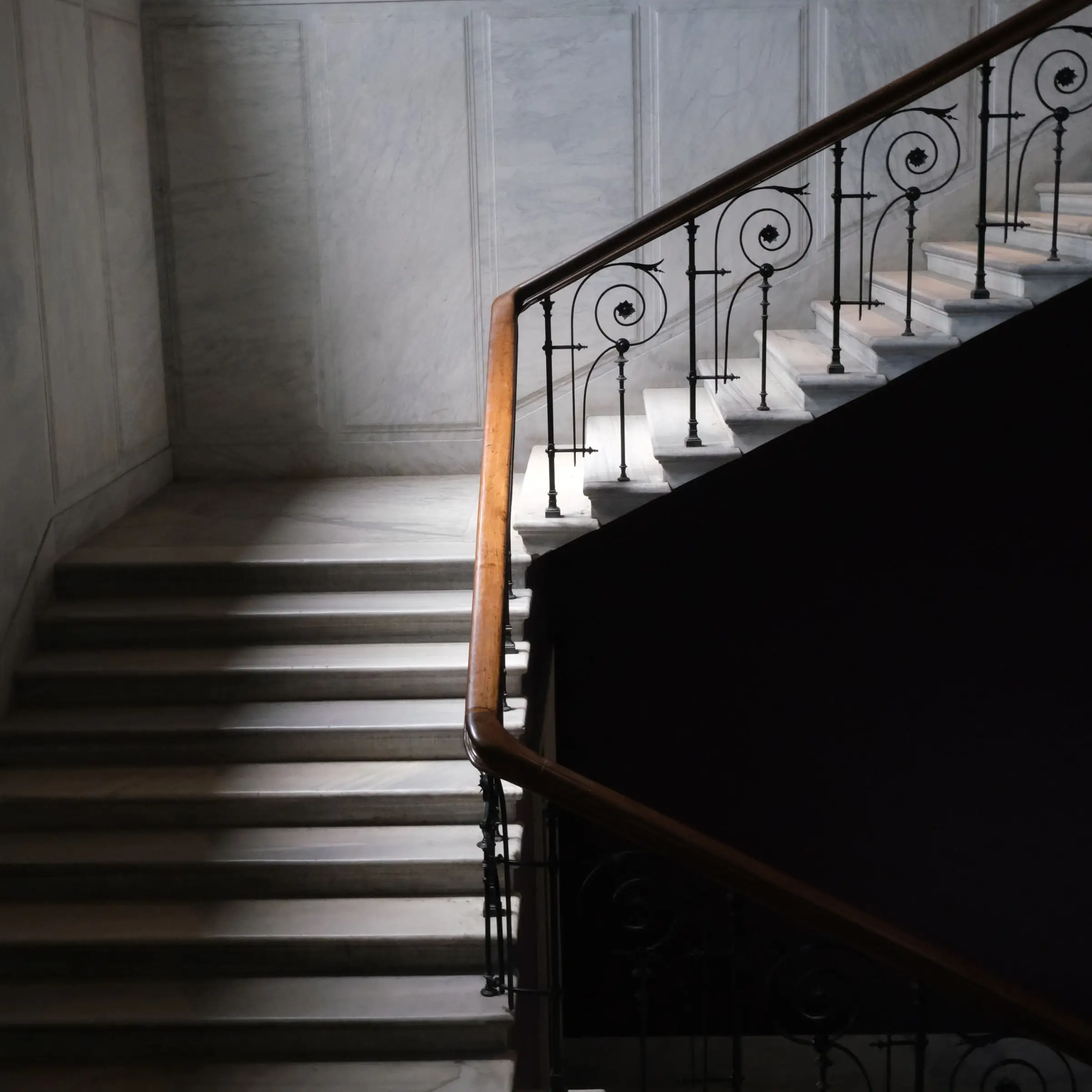 Escalier en marbre clair aux marches massives à nez droit, rampe en fer forgé, lumière latérale froide