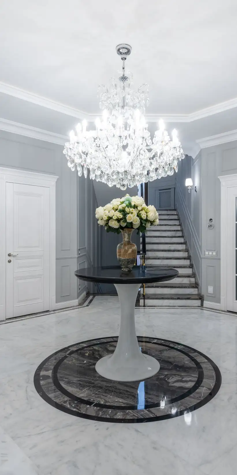 Hall d’entrée au sol de marbre clair poli avec médaillon circulaire sombre, escalier au fond, portes blanches