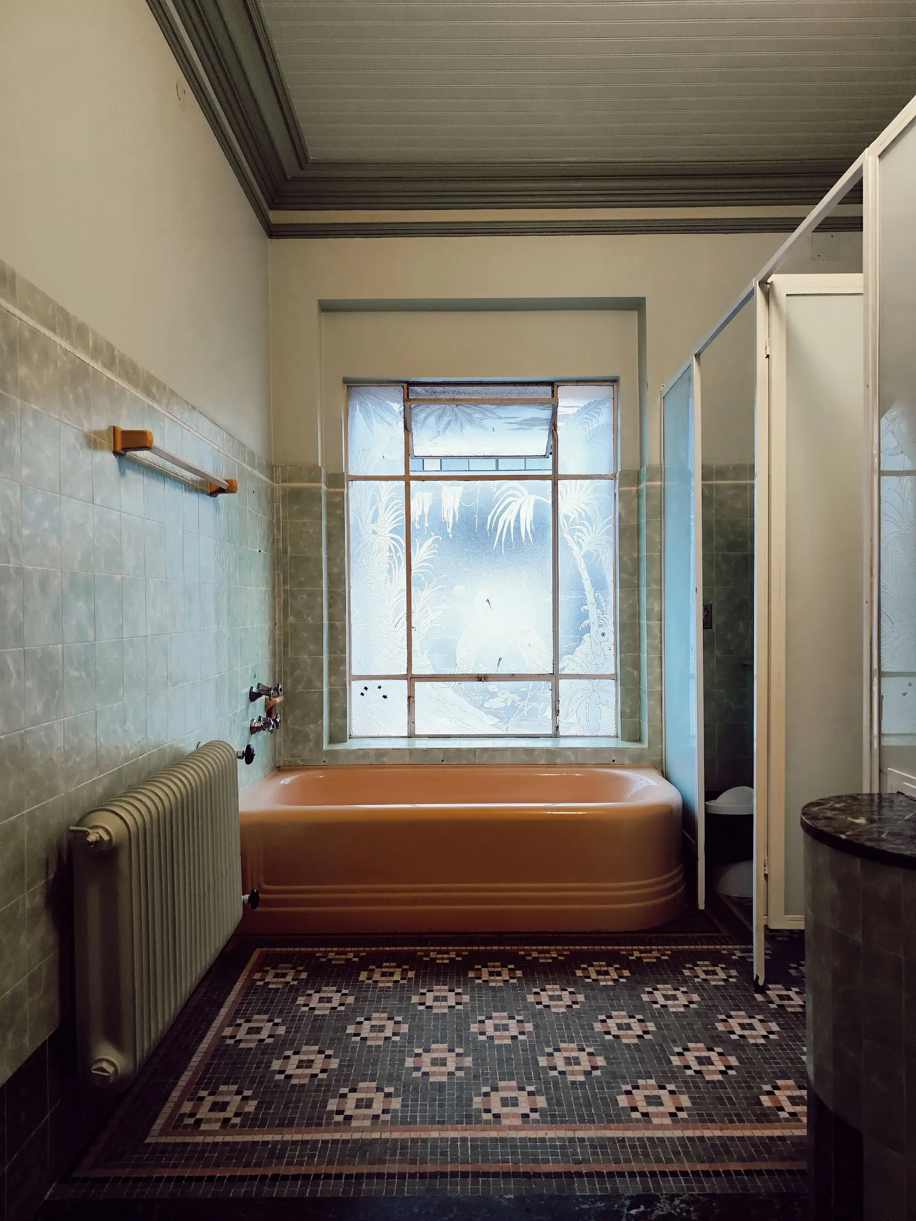Salle de bain ancienne : baignoire brune encastrée, faïence pâle, sol en mosaïque, radiateur sous une fenêtre