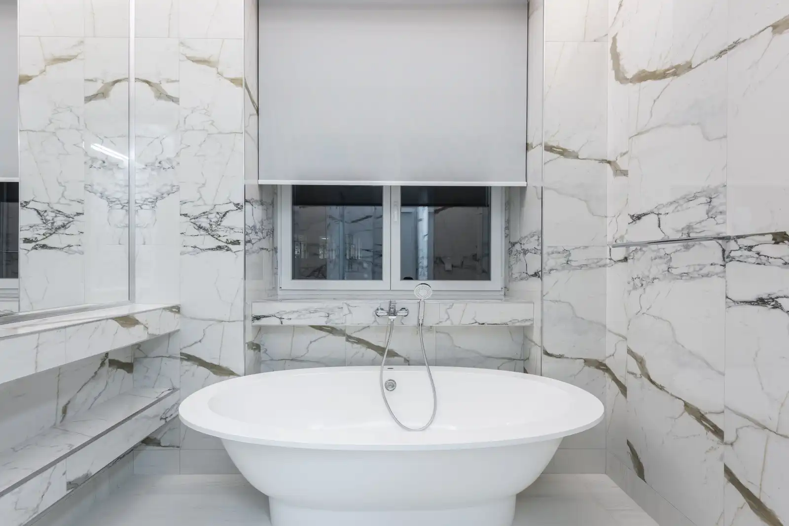 Salle de bain finie en marbre blanc veiné de gris et d’or : murs en grandes dalles, baignoire îlot sous une fenêtre centrée, plan de toilette en marbre, lumière du jour diffuse
