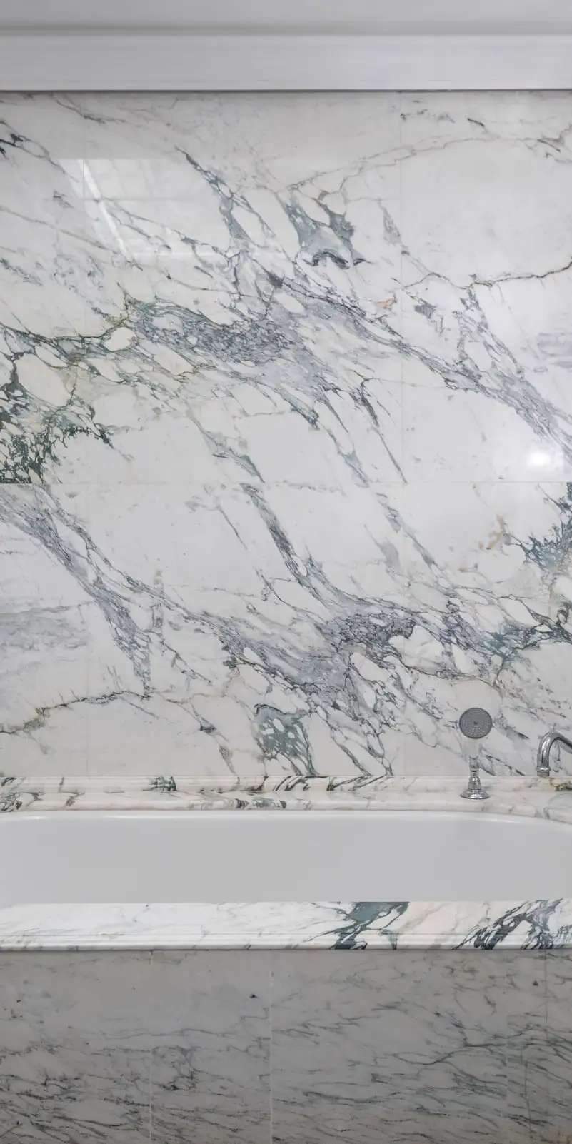 Mur de salle de bain en marbre blanc aux larges veines gris-vert posé en livre ouvert, baignoire habillée du même marbre, sèche-serviettes chromé