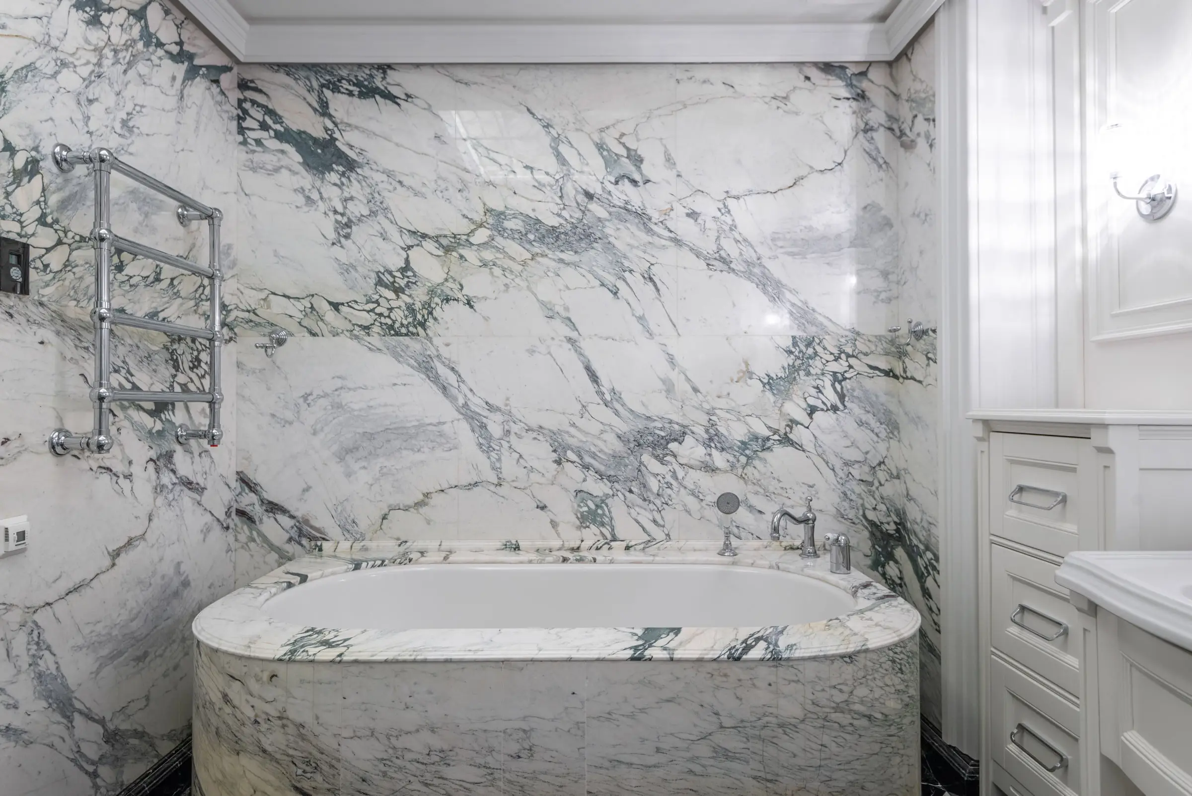 Mur de salle de bain en marbre blanc aux larges veines gris-vert posé en livre ouvert, baignoire habillée du même marbre, sèche-serviettes chromé