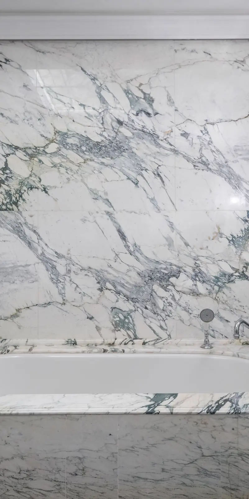 Mur de salle de bain en marbre blanc aux larges veines gris-vert posé en livre ouvert, baignoire habillée du même marbre, sèche-serviettes chromé